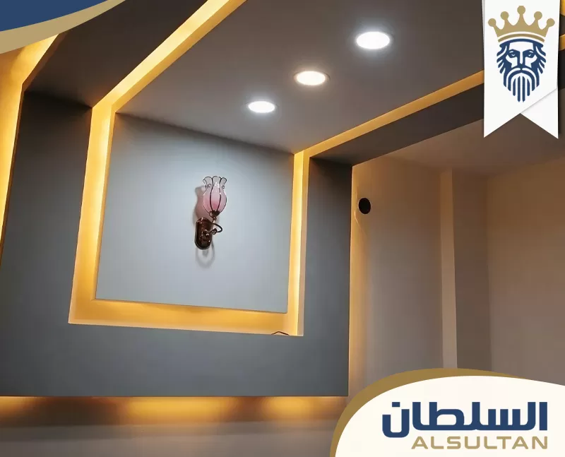 ديكور جبس بورد للجدار والسقف، بتجاويف هندسية وإضاءة مخفية دافئة، مع سبوت لايت سقفي حديث داخل الغرفة.